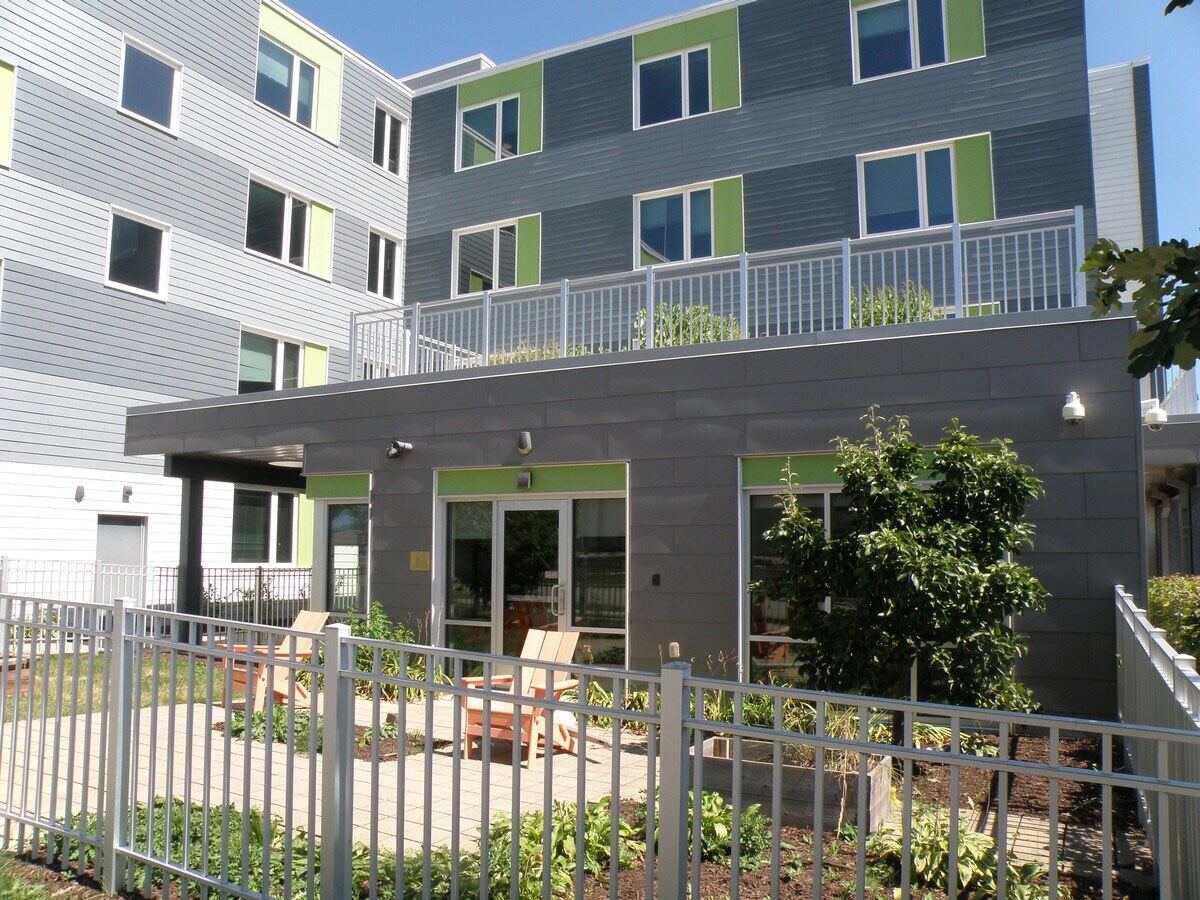 Courtyard of a four-story residential building with green window accents, a fenced garden and Adirondack chairs