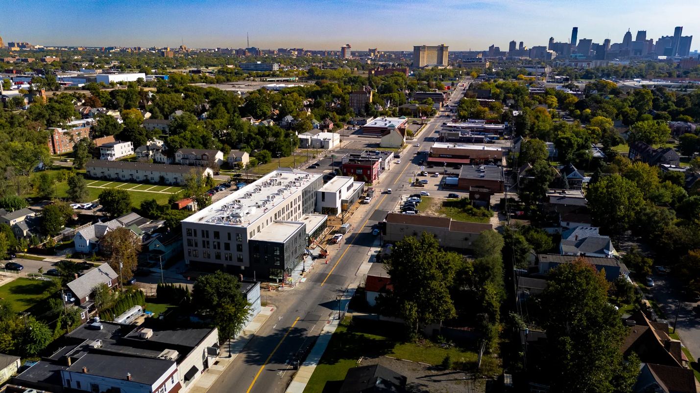 Aerial view of a new three-story building along a neighborhood main street in Detroit, with the downtown skyline in the distance